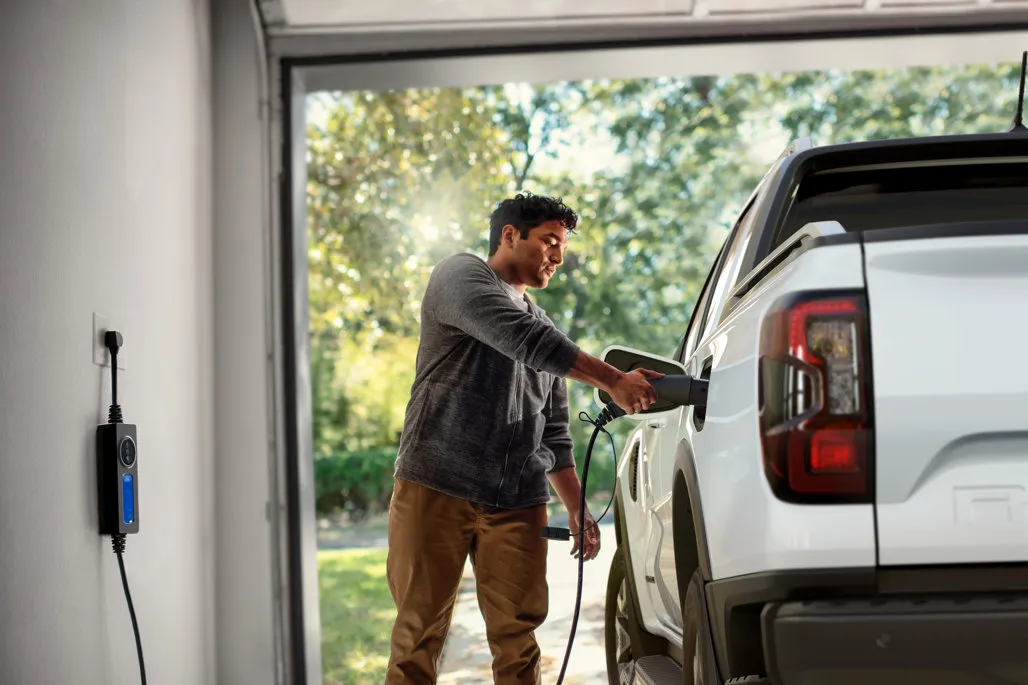 A man wearing a grey hoodie plugs a charging cable into the port of a white electric pickup truck parked inside a residential garage.