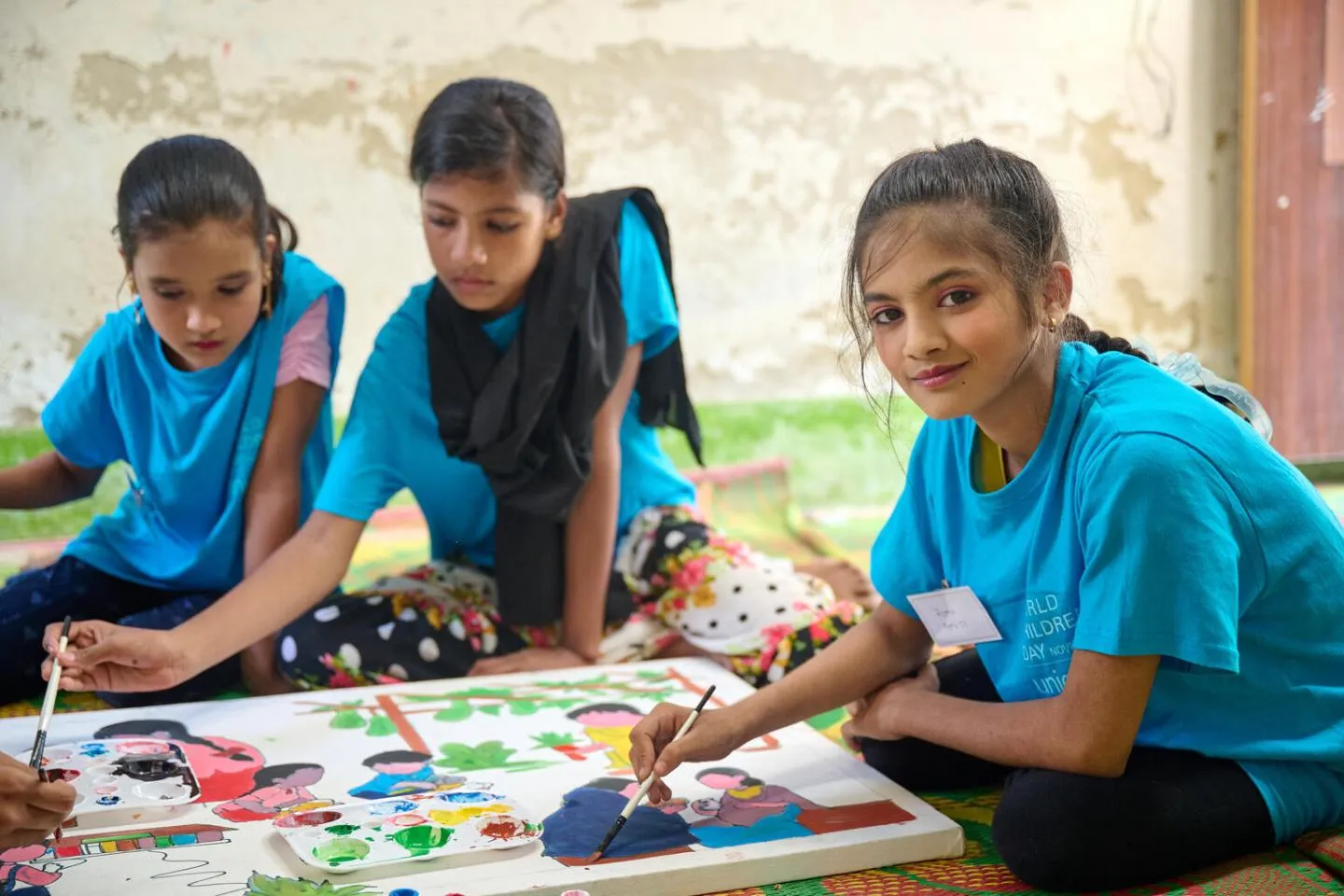 Three young girls in blue t-shirts sitting on a floor mat, collaboratively painting a colorful mural with brushes and palettes, with one girl smiling directly at the camera during a World Children's Day event.