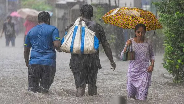 Three people wading through a heavily flooded street during a downpour, with a woman holding a yellow patterned umbrella and a man carrying a large striped bag on his shoulder.