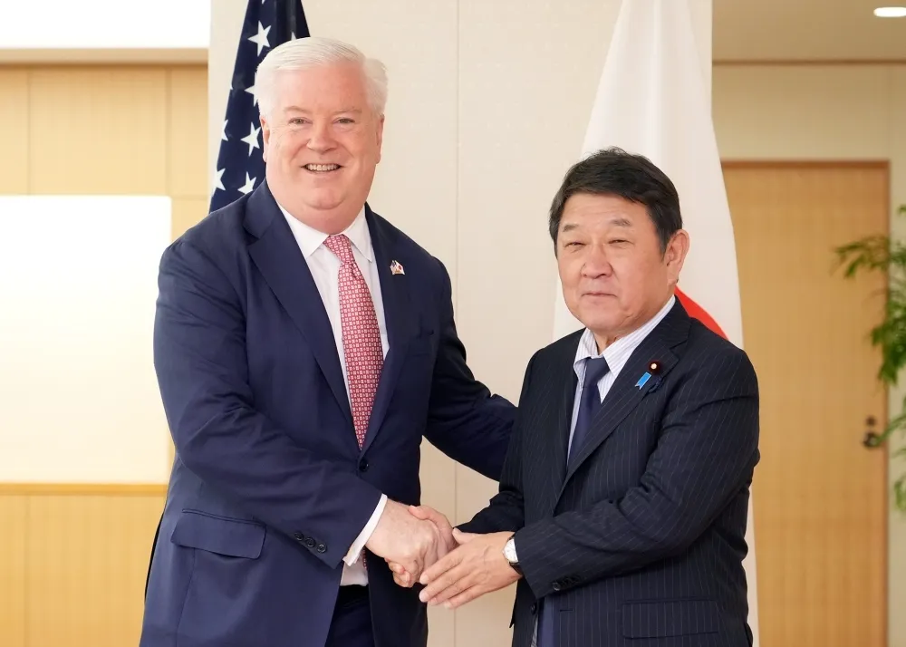 U.S. Ambassador George Glass shakes hands with Japanese Foreign Minister Toshimitsu Motegi in Tokyo, flanked by U.S. and Japanese flags.