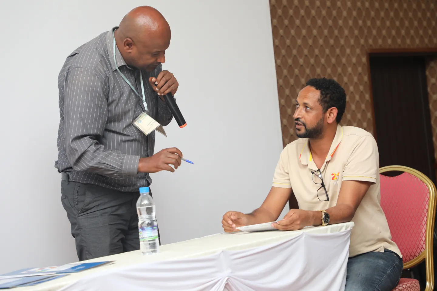 A standing man in a grey striped shirt holds a microphone while conducting a role-play skills assessment with Ashenafi Semerga, who sits at a white-clothed table during an ILO workshop in Hawassa, Ethiopia.