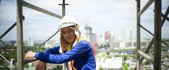 A construction worker wearing a white hard hat, sun protection cloth, and a blue long-sleeve shirt leans on metal scaffolding high above a city skyline during the day.