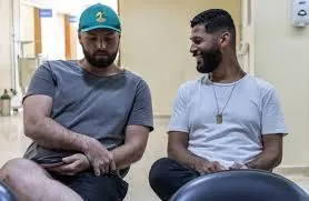 Two men sitting side-by-side in a medical clinic waiting area; one man wears a grey t-shirt and green baseball cap while looking down, and the other wears a white t-shirt and smiles at him.