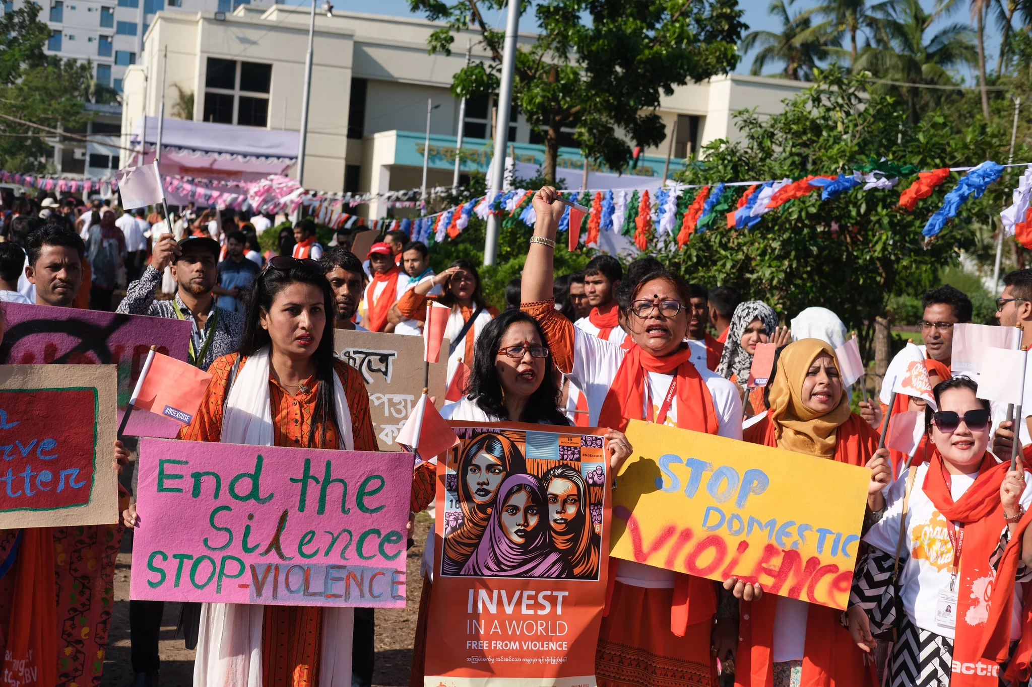Activists and women march outdoors holding colorful signs reading "End the Silence Stop Violence," "Stop Domestic Violence," and "Invest in a World Free From Violence," while wearing orange scarves to protest gender-based violence.