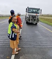 A female traffic officer wearing a high-visibility reflective vest and orange dress stands on a wet roadside, looking back at the camera, while two colleagues and a large white truck pass on the road behind her during the Gauteng Points Person of the Future launch.