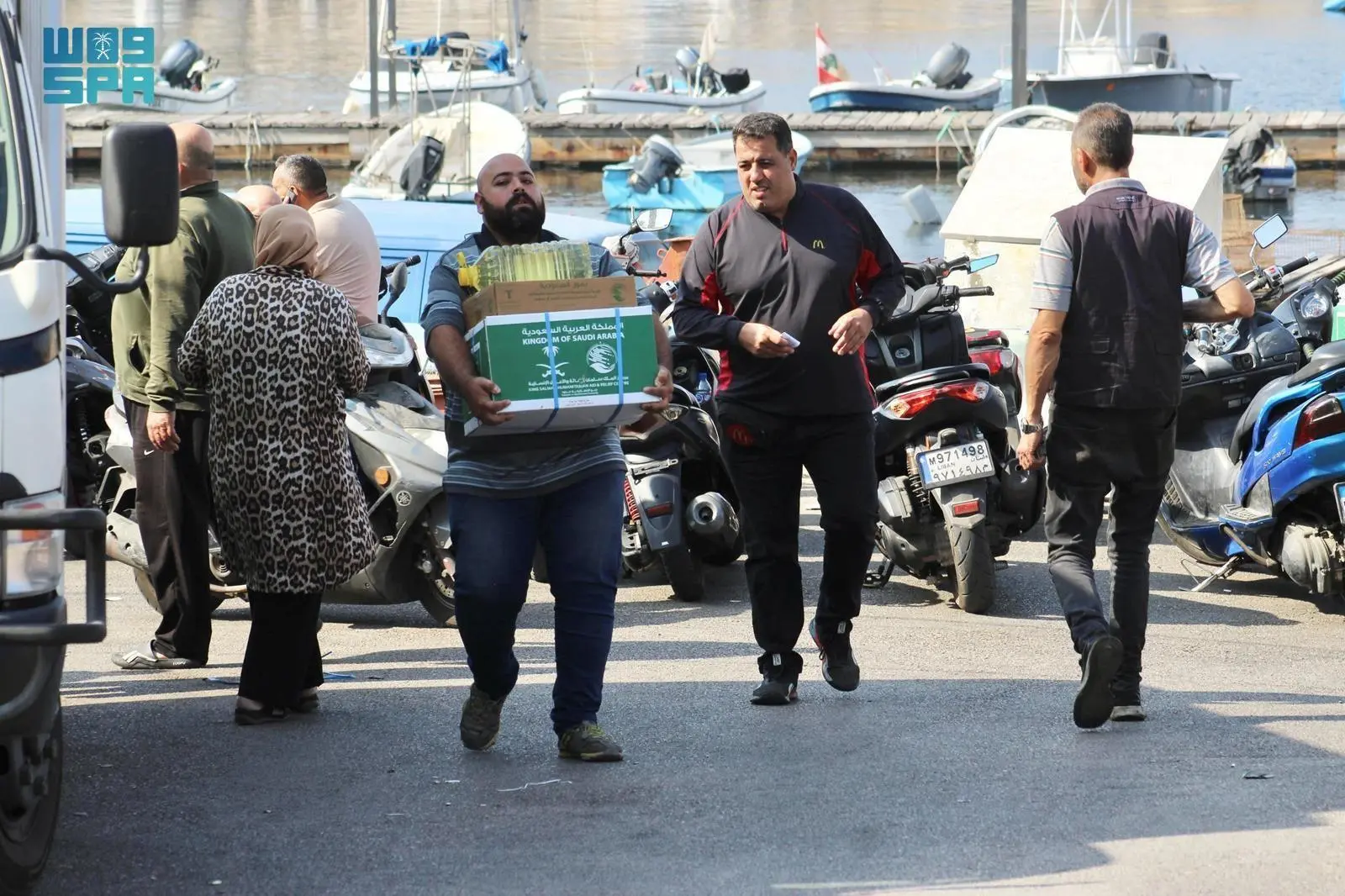 A man carries a green aid box bearing the King Salman Humanitarian Aid and Relief Center logo and a bottle of cooking oil near a harbor in Naameh, Lebanon, during a distribution event.