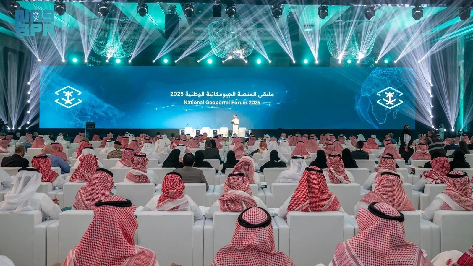 Attendees in traditional Saudi attire watch a presentation on a large blue stage screen reading National Geoportal Forum 2025 during the event organized by GEOSA in Riyadh.