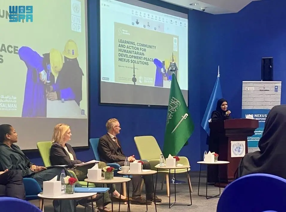 A female speaker stands at a podium addressing attendees during a KSrelief and UNDP workshop in Riyadh, with a large screen displaying 'Nexus Academy' and a Saudi Arabian flag nearby. Three panelists sit on stage listening to the presentation.