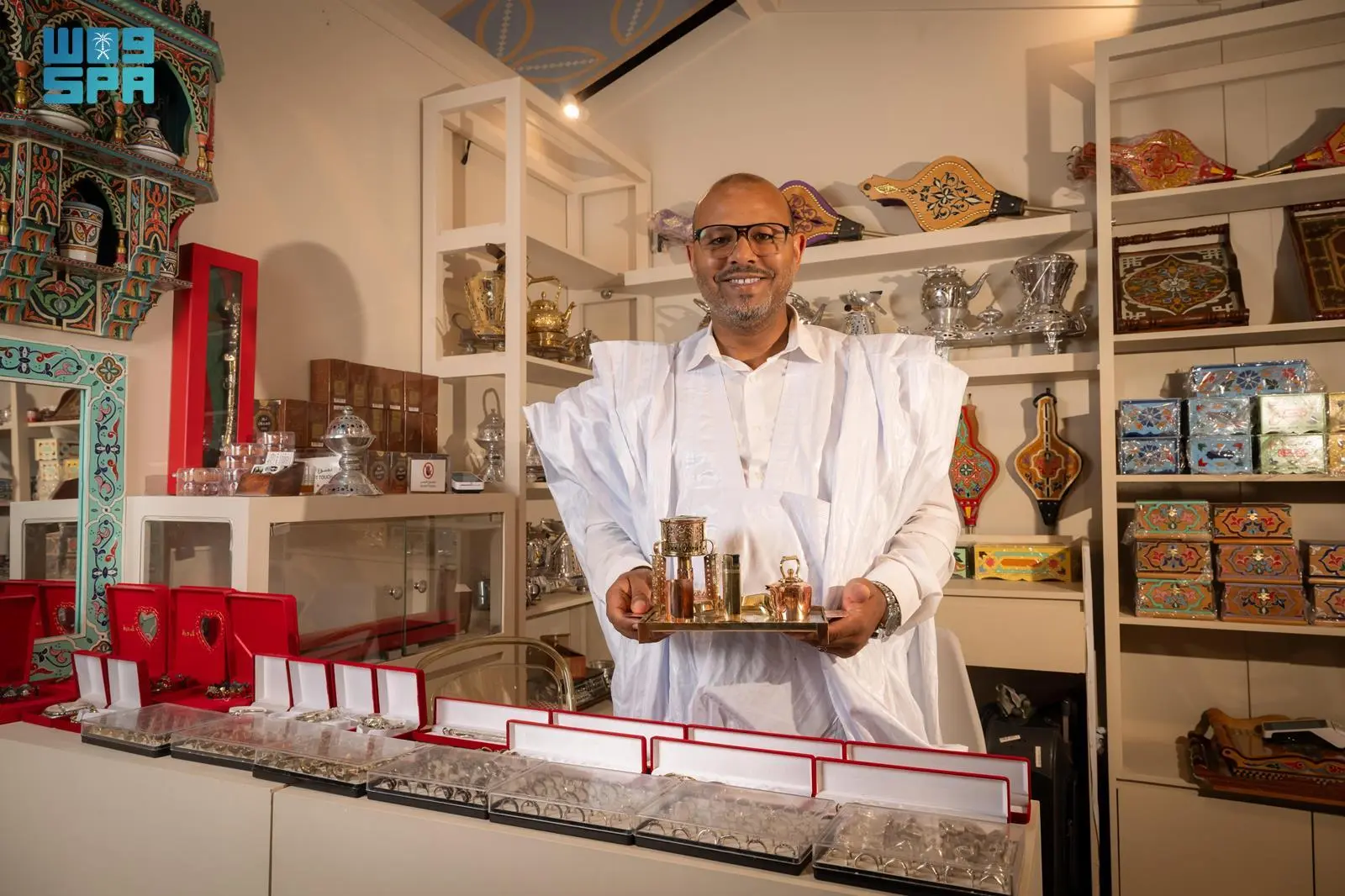 Moroccan artisan Andislem Yara, wearing traditional white attire, holds a copper tea set on a tray while standing inside his booth filled with silver handicrafts and decorative boxes at the Saudi International Handicrafts Week.