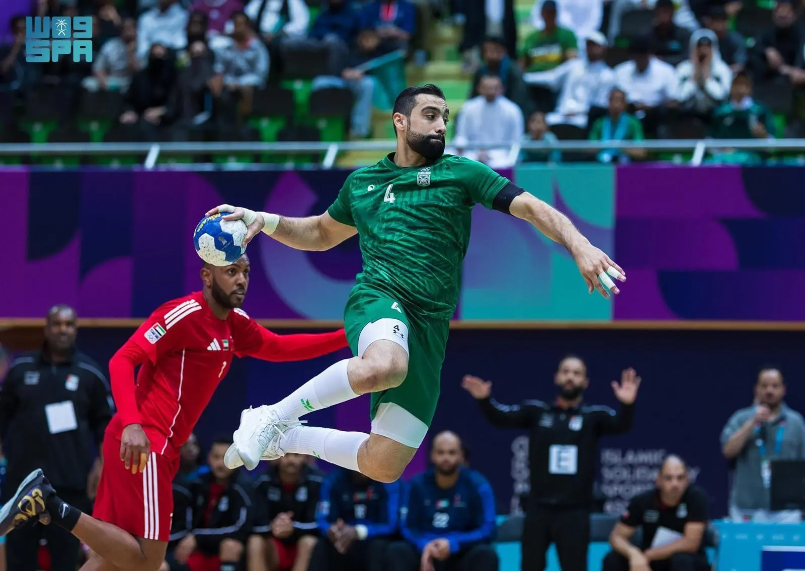 A Saudi national team handball player in a green number 4 jersey executes a high jump shot while being defended by a UAE player in red during the 6th Islamic Solidarity Games semifinal.