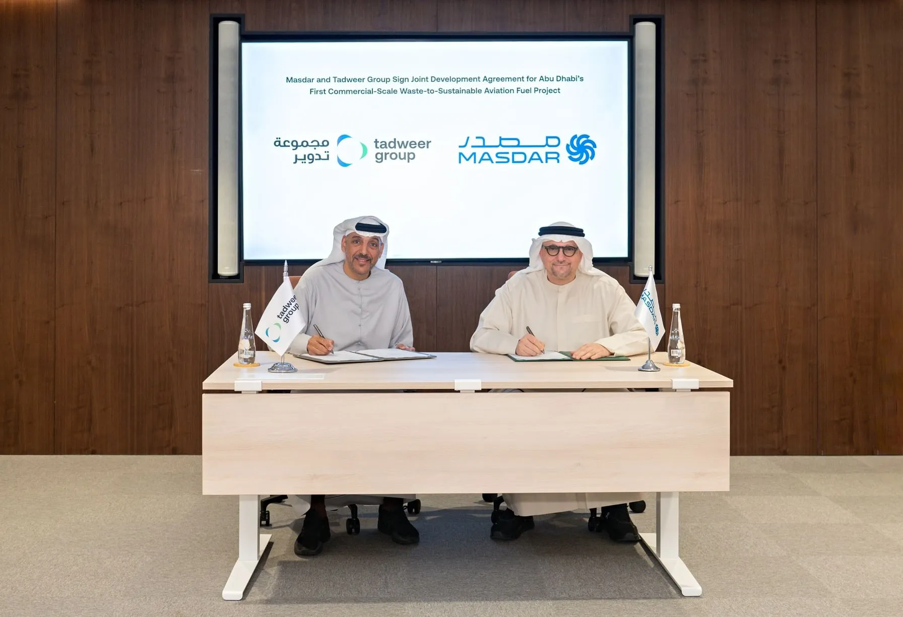 Tadweer Group MD Ali Al Dhaheri and Masdar CEO Mohamed Jameel Al Ramahi signing a joint development agreement for a waste-to-sustainable aviation fuel project in Abu Dhabi, seated at a table with a digital screen displaying company logos in the background. 