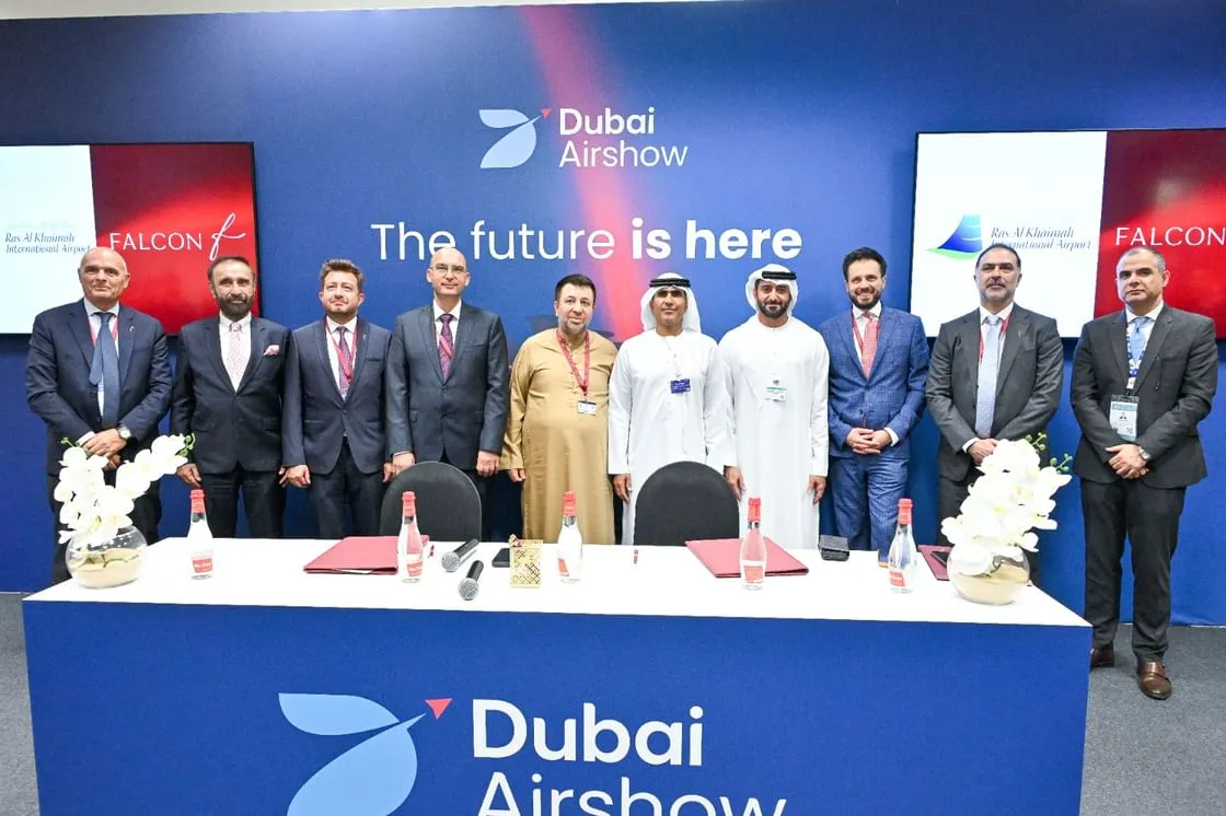 officials from Ras Al Khaimah International Airport and Falcon Executive Aviation posing for a group photo after signing an agreement at the Dubai Airshow, with screens displaying the partners' logos in the background.