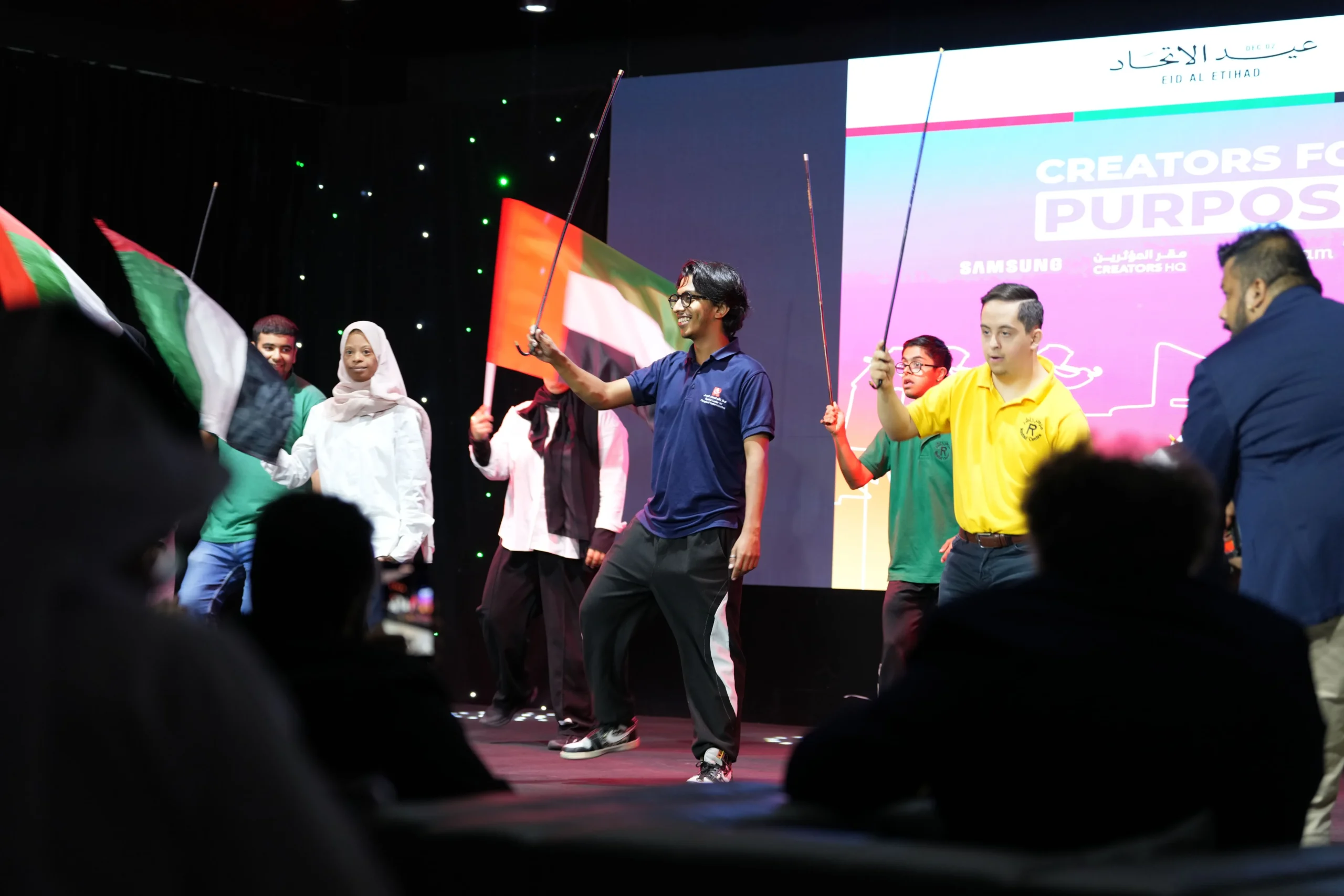 A group of young men and women performing on stage holding UAE flags and thin canes. A man in a blue polo shirt smiles center stage while holding a cane, next to a man in a yellow shirt. A large digital screen in the background displays the text 'CREATORS FOR PURPOSE' and 'EID AL ETIHAD' alongside the Samsung logo.