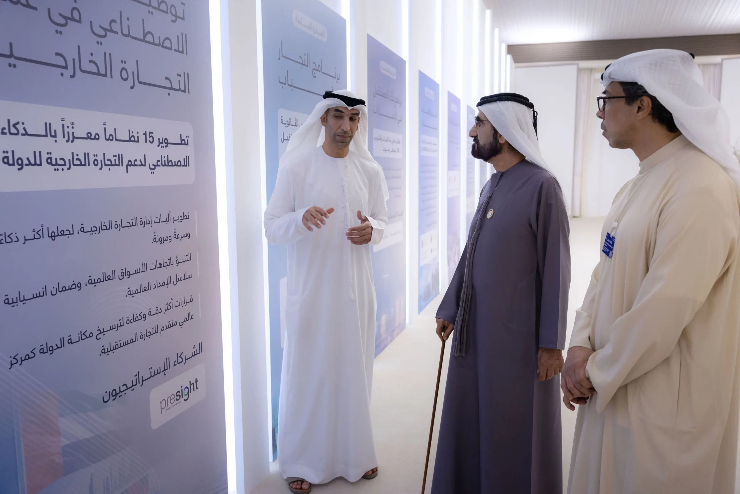 Sheikh Mohammed bin Rashid Al Maktoum listens to Dr. Thani bin Ahmed Al Zeyoudi explaining the new UAE Global Centre of Trade strategy in front of display panels detailing AI integration in foreign trade.