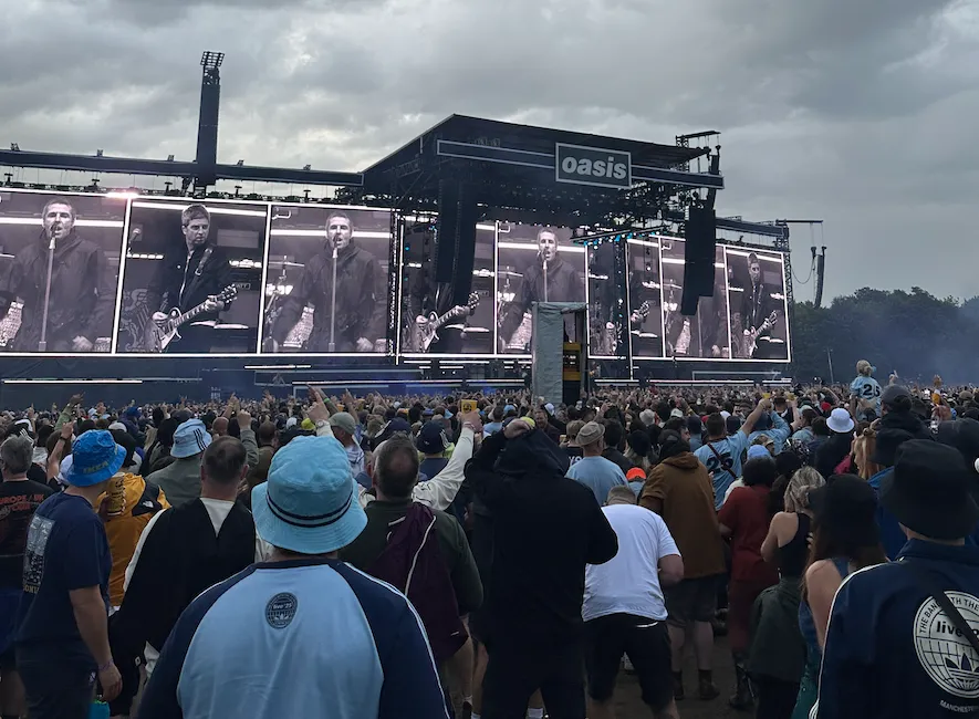 A large crowd of music fans stands before a massive outdoor stage structure featuring the Oasis logo, with band members performing on large vertical screens under a cloudy sky.