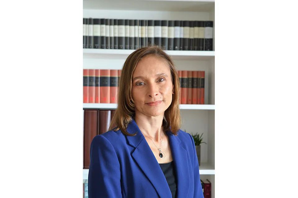 Portrait of UK Ambassador Corinne Kitsell wearing a royal blue blazer and silver necklace standing in front of a white bookshelf filled with bound volumes.