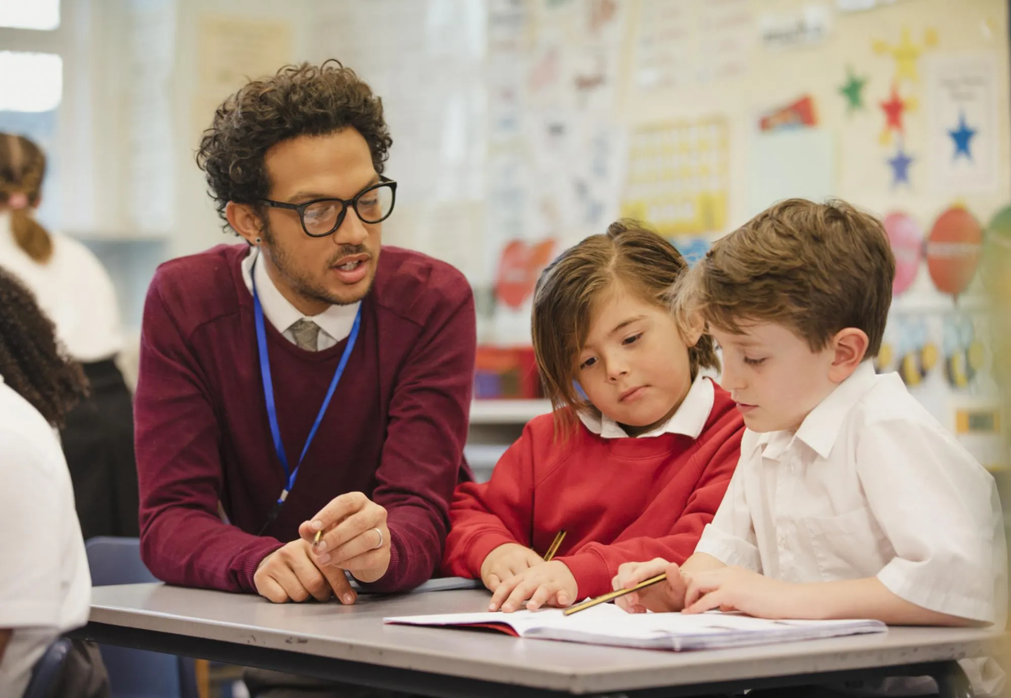 A male teacher wearing glasses and a burgundy sweater leans over a desk to assist a young girl and boy with their written schoolwork in a classroom setting.