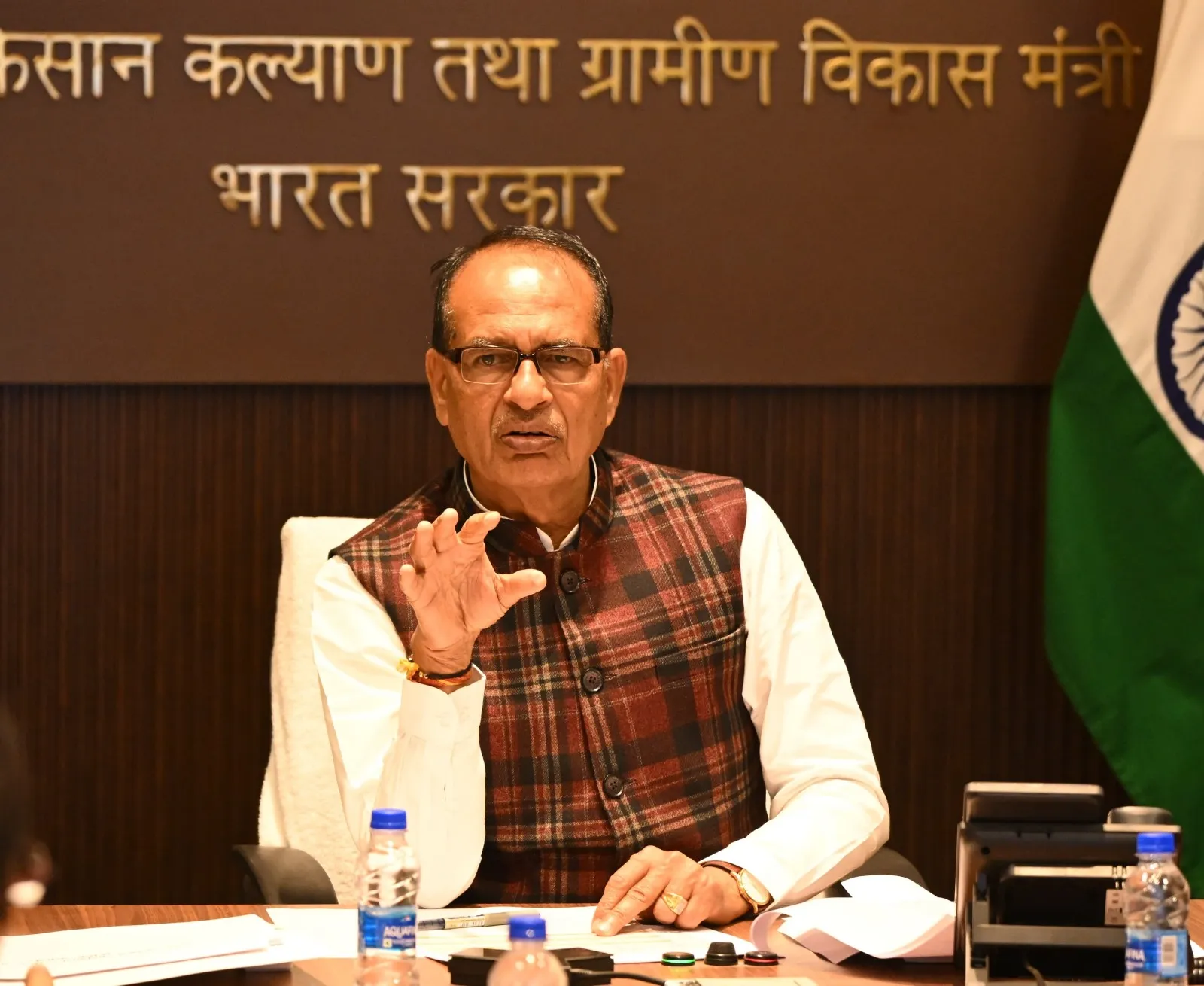Union Agriculture Minister Shivraj Singh Chouhan wearing a maroon checkered vest and glasses, gesturing with his right hand while speaking at a desk with the Ministry of Agriculture nameplate in Hindi in the background.