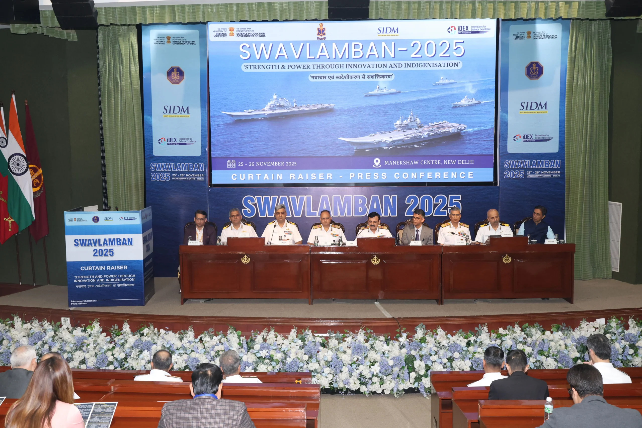 Vice Admiral Sanjay Vatsayan and Indian Navy officials seated at a long dais during the Swavlamban 2025 curtain raiser press conference in New Delhi, with a large screen displaying aircraft carriers in the background.
