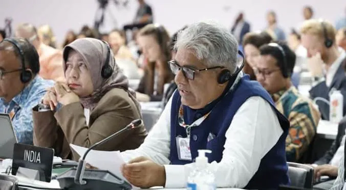 Union Minister Bhupender Yadav representing India at the CoP30 conference in Belém, Brazil, wearing headphones and a blue vest while seated at a desk with an 'INDIA' nameplate.