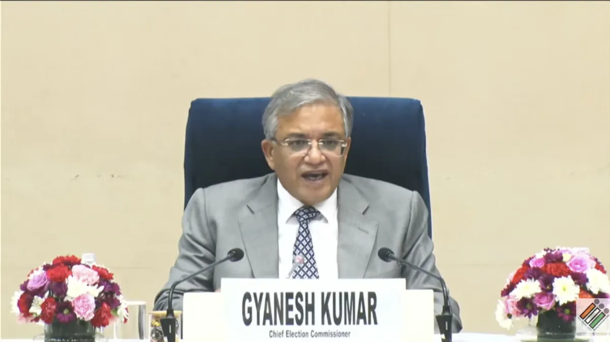 Chief Election Commissioner Gyanesh Kumar sitting at a desk with a nameplate reading "GYANESH KUMAR" and microphones, addressing a press conference with the Election Commission of India logo visible in the corner.