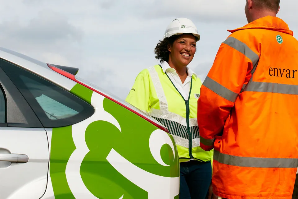 A smiling Environment Agency officer wearing a white hard hat and yellow high-visibility vest converses with a worker in an orange Envar jacket next to a branded agency vehicle.