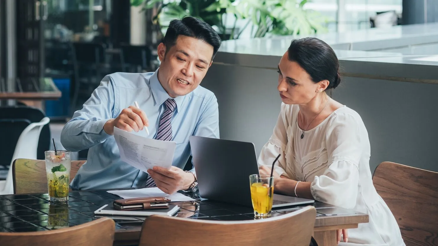 Asian male professional explaining document details to a woman using a laptop during a business meeting in a cafe setting.
