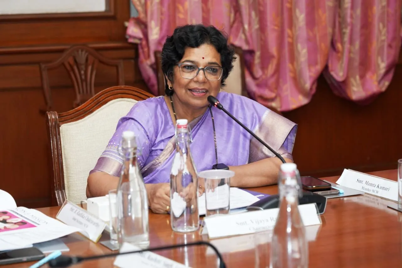 Smt. Vijaya Rahatkar, Chairperson of the National Commission for Women (NCW), wearing a purple saree and glasses, speaks into a microphone while presiding over a formal meeting at the NCW headquarters in New Delhi; a nameplate identifying Member Smt. Mamta Kumari is also visible.