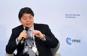 Japanese government official in a dark suit speaking into a microphone at the Munich Security Conference with MSC branding in the background