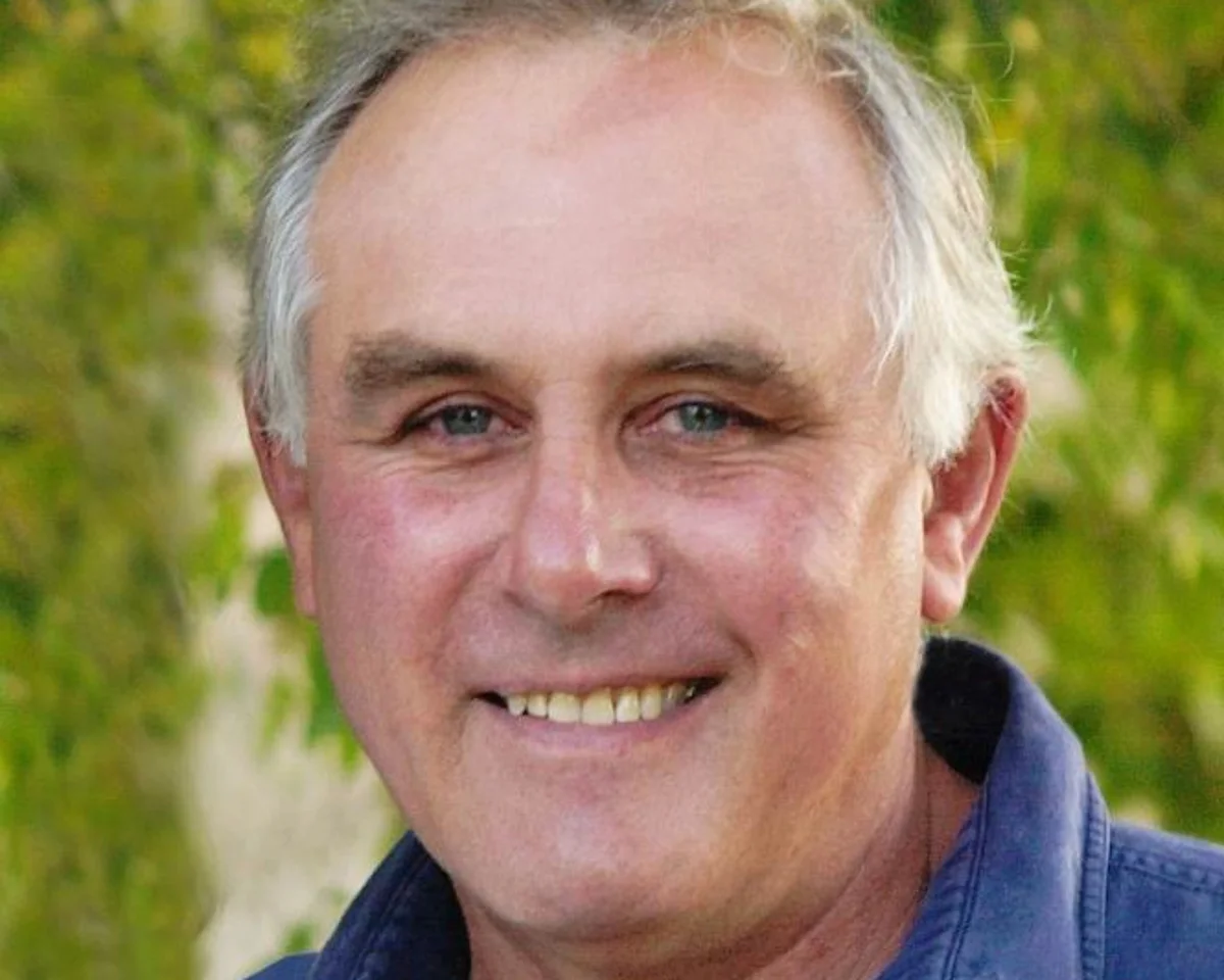 Headshot of Bruce Haigh, former Australian diplomat and anti-apartheid activist, smiling in a blue shirt against a blurred green natural background.