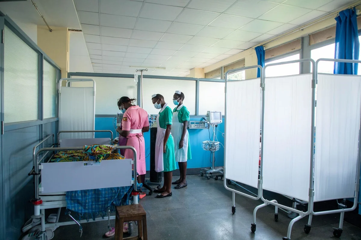 Three nurses in distinct pink and green uniforms attend to a patient in a hospital bed surrounded by privacy screens and medical equipment in a ward in Uganda or Malawi