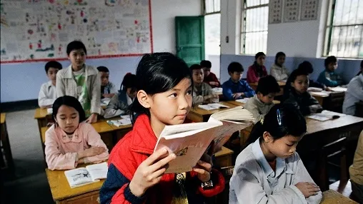 A young female student in a red jacket stands holding an open book in a classroom, while other students sit at desks in the background.