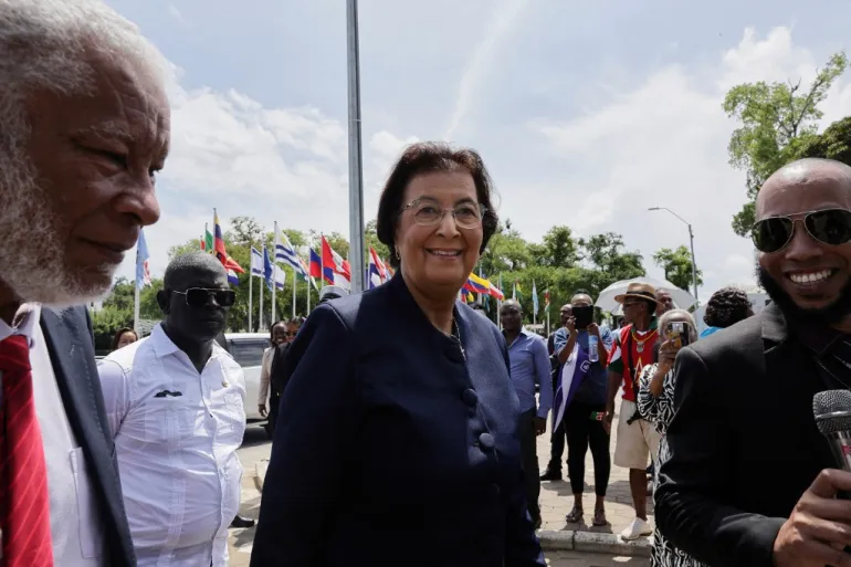 A female official in a navy blue suit and glasses smiles while speaking to a reporter holding a microphone, flanked by a man with a white beard and red tie, with international flags and trees in the background in Paramaribo.