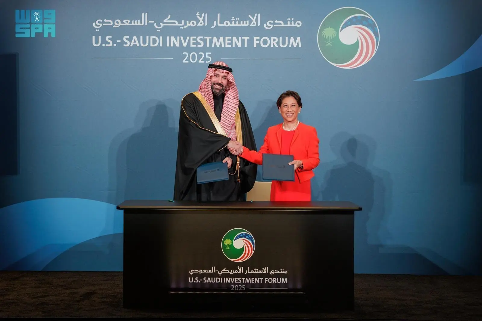 A representative from the Saudi National Development Fund and a State Street executive shake hands while holding signed agreement folders at the U.S.-Saudi Investment Forum 2025 podium.