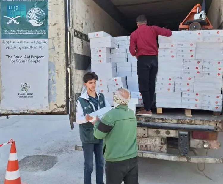 A KSrelief aid worker hands a carton of dates to a man in a green jacket while another worker stacks white boxes inside a truck branded with the Saudi Aid Project for Syrian People logo.