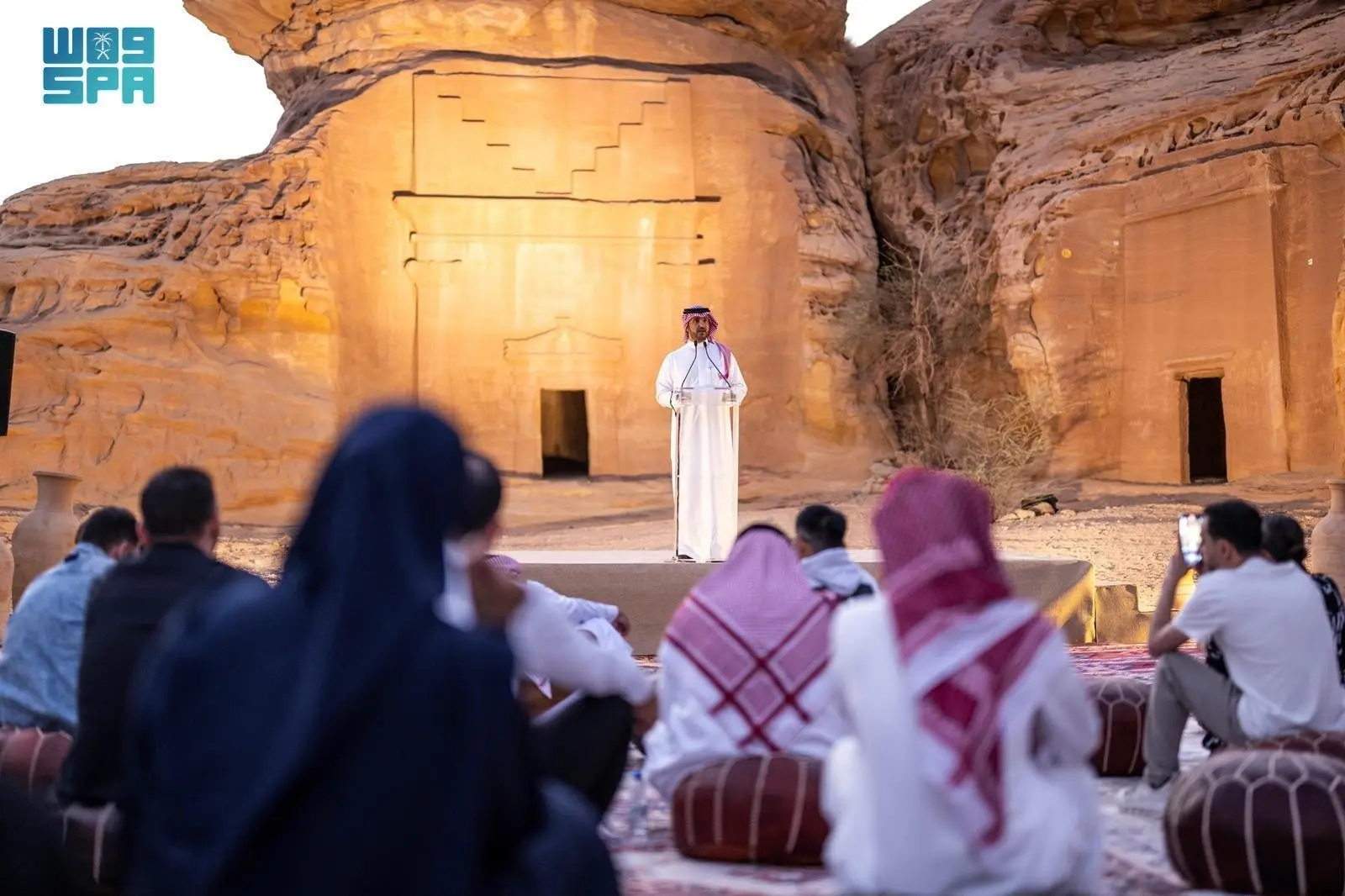 Dr. Abdulrahman Alsuhaibani speaking at a podium during the Ancient Kingdoms Festival opening ceremony in front of a rock-cut tomb at Al Hijr, AlUla.