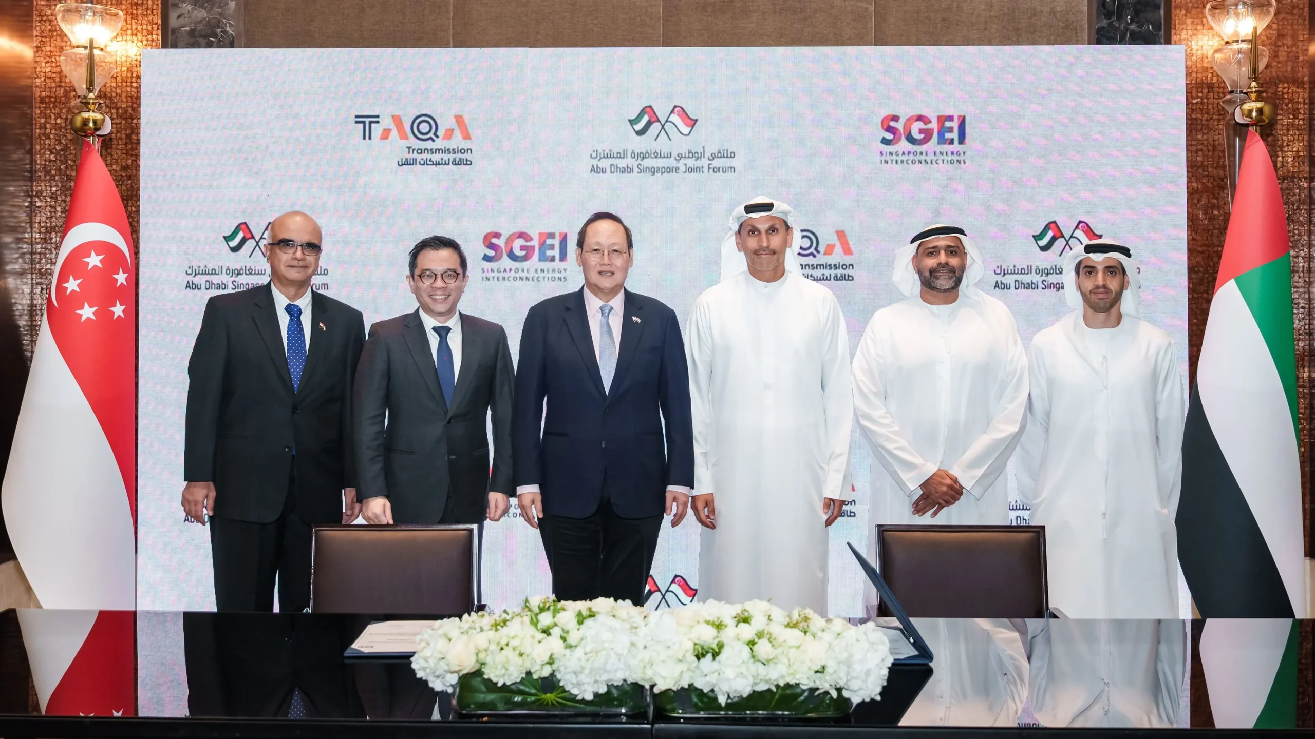 Delegates from the UAE and Singapore standing in a row for a group photo during the 16th Abu Dhabi–Singapore Joint Forum, flanked by national flags and logos of TAQA and SGEI.