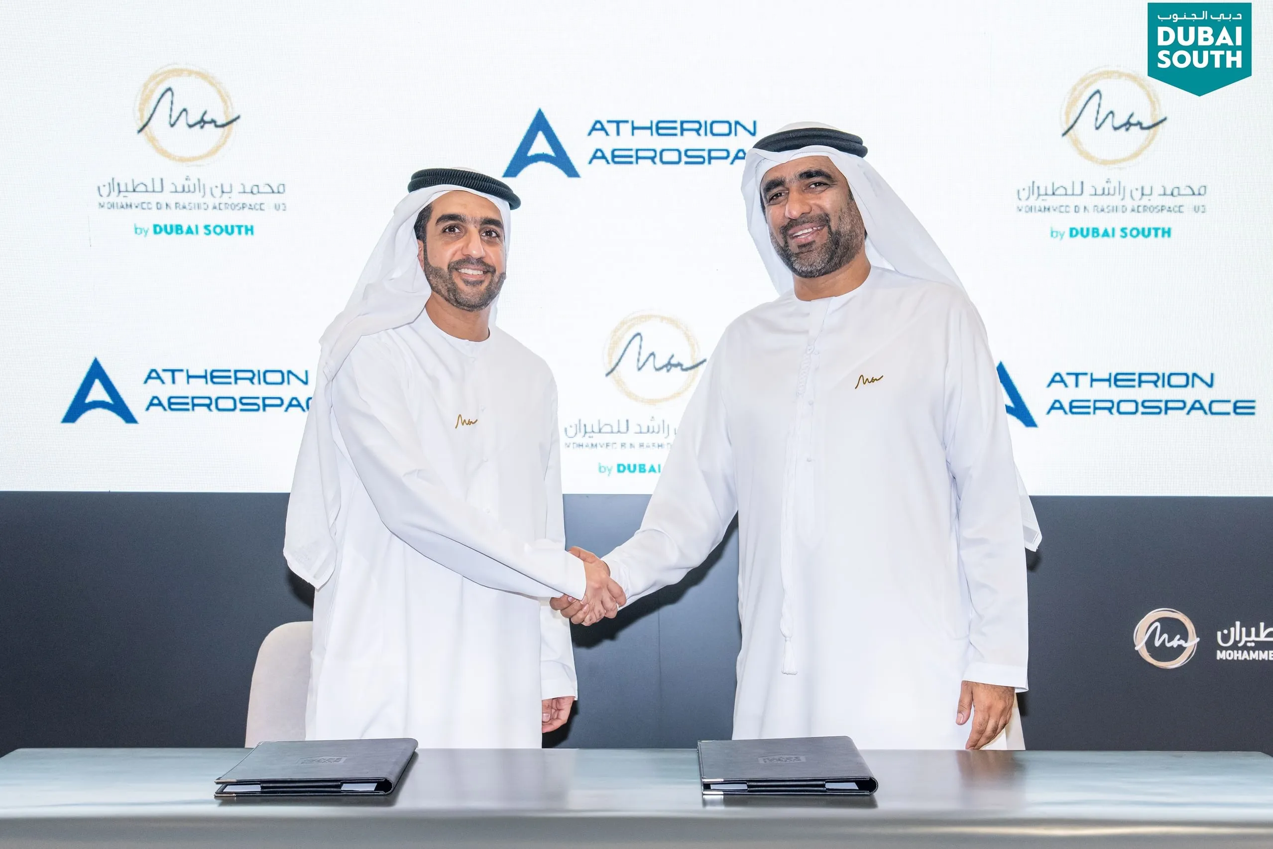 Tahnoon Saif and Sheikh Sultan Majid Hamad Binsaqar Alqasimi shaking hands after signing an agreement between MBRAH and Atherion Aerospace in front of a branded backdrop.