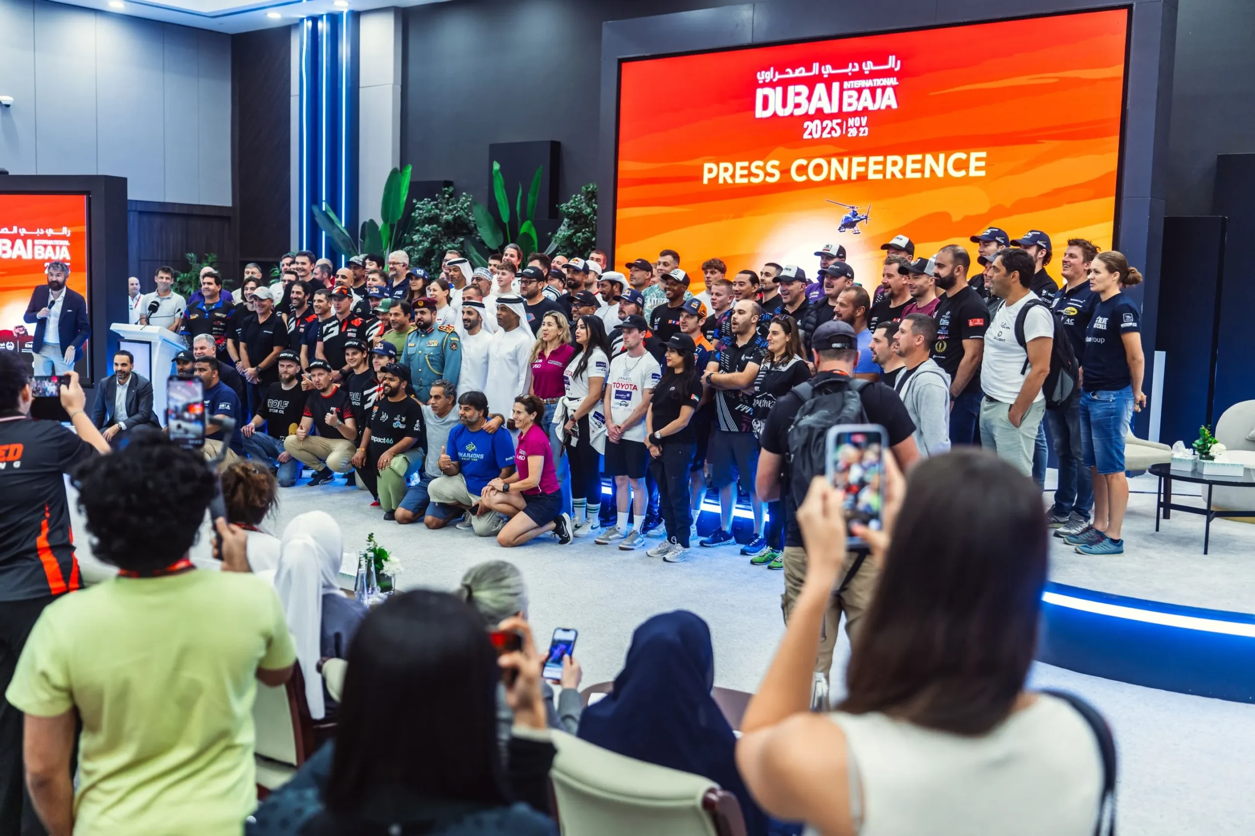 A large group of international off-road rally drivers, team members, and officials pose on a tiered stage during the Dubai International Baja 2025 press conference, backed by a large orange screen displaying the event logo and dates, while audience members in the foreground take photos with smartphones.