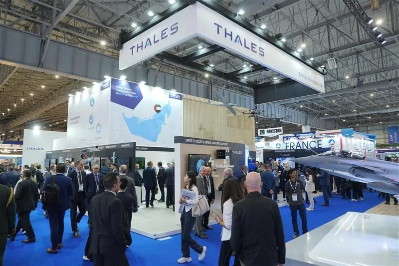 A wide shot of the Thales exhibition stand at the Dubai Airshow 2025, featuring a large overhead banner with the company logo, a wall graphic displaying a map of the UAE, suspended drone models, and visitors interacting with defense and aviation displays.