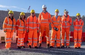 Seven construction project leaders wearing orange high-visibility jackets and hard hats stand in a line in front of a large mound of recycled concrete aggregate at the Sizewell site.