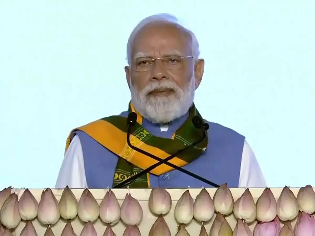 Prime Minister Narendra Modi addressing an event, wearing a light blue vest and a yellow-green traditional scarf, standing behind a podium decorated with lotus flowers.