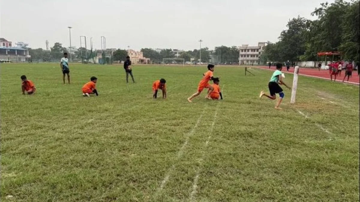 Young athletes competing in a game of Kho Kho on a grassy field during the Kashi MP Sports Competition, featuring players kneeling in a line while a runner turns around a pole.
