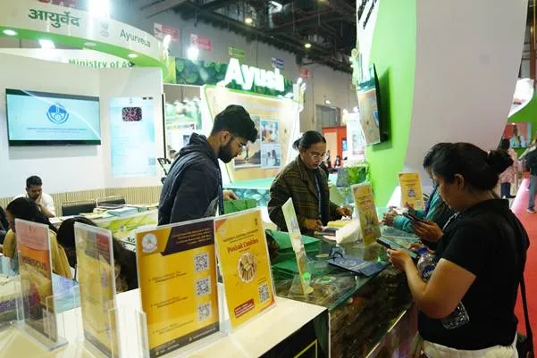 Visitors interacting with staff at the Ministry of Ayush pavilion during IITF 2025, featuring displays of Ayurvedic products like Poshak Cookies, informational brochures, and large green Ayush branding.