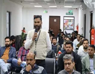 A participant speaking into a microphone during the Telecom Outreach Meet organized by the Office of Principal Controller of Communication Accounts, Delhi, with other attendees seated in rows behind him.