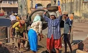 A female construction worker carries a metal basin on her head while male and female laborers work with a concrete mixer and shovels at a construction site in India.