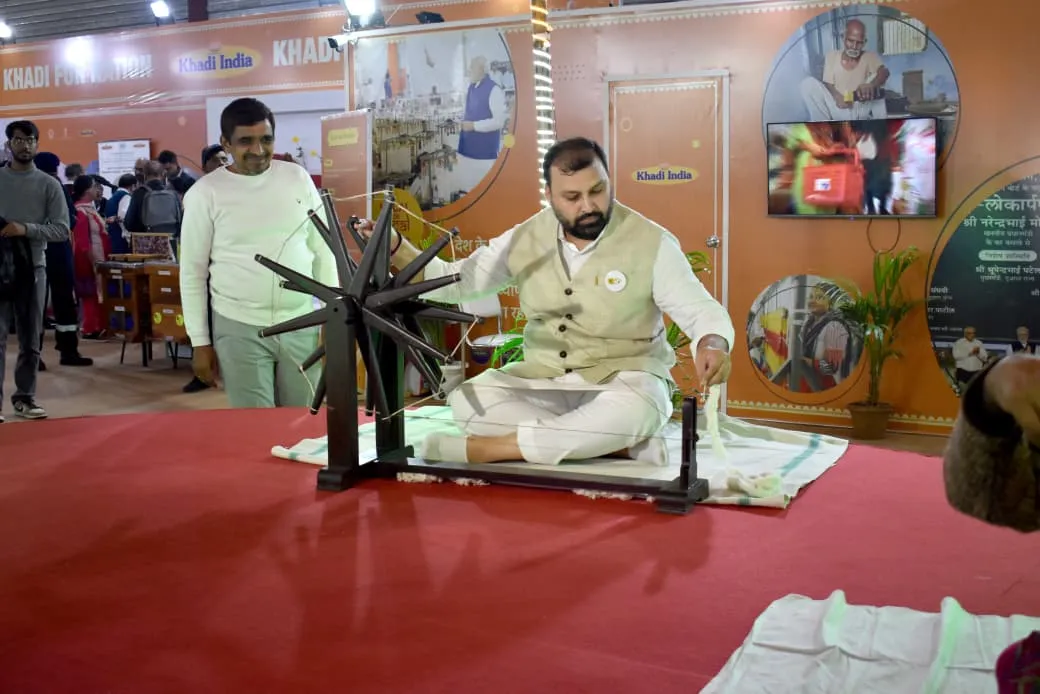 KVIC Chairman Manoj Kumar sitting cross-legged on a red platform spinning a large traditional black charkha wheel at the Khadi India Pavilion during IITF 2025.