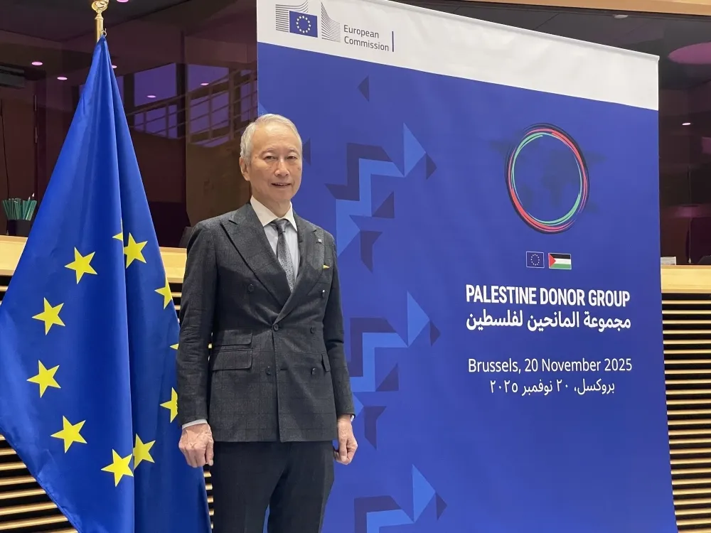 Ambassador Okubo Takeshi standing in a grey suit next to a European Union flag and a blue banner for the Palestine Donor Group meeting in Brussels, dated November 20, 2025.