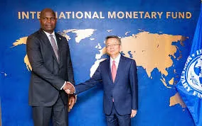Bank of Mozambique Governor Rogério Zandamela (left) shakes hands with a high-ranking IMF official in front of a blue wall featuring the International Monetary Fund logo and a gold world map.
