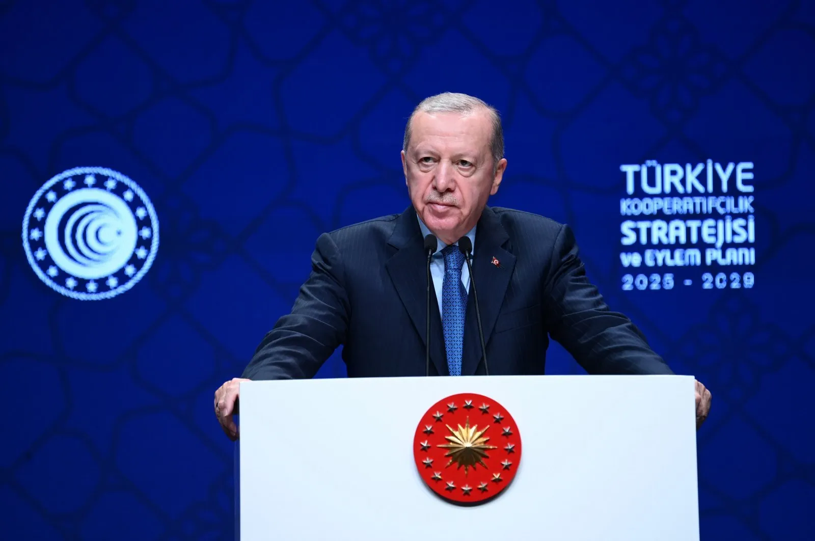 Turkish President Recep Tayyip Erdoğan delivers a speech at the Turkey Cooperatives Strategy and Action Plan 2025-2029 launch, standing behind a podium with the presidential seal against a blue patterned background.